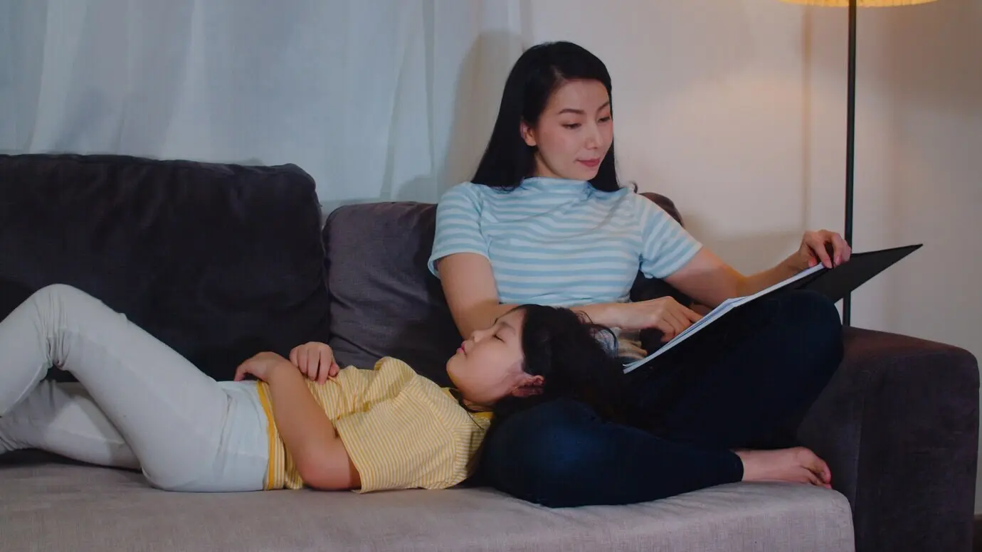 Eine junge asiatische Mutter liest ihrer Tochter zu Hause Märchen vor. Eine glückliche chinesische Familie entspannt sich mit einem Teenager-Mädchen, das beim Zuhören der Geschichten nachts im Schlafzimmer eines modernen Hauses im Bett schläft.